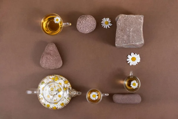 Chamomile tea. Cups of herbal tea, transparent teapot with camomile flowers and stones on brown background. Calming drink concept. Trendy still life.