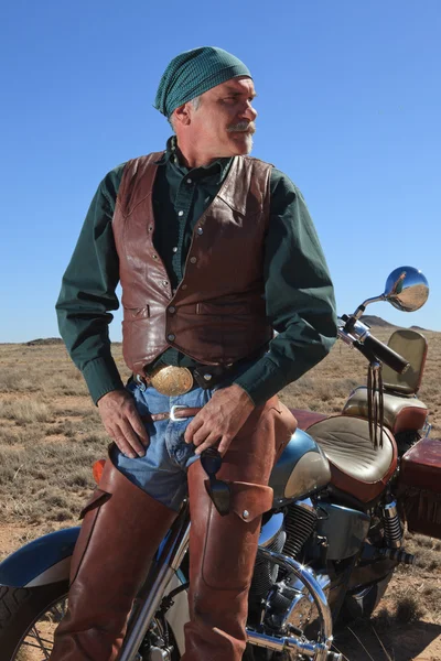 Bell'uomo anziano in pensione nel deserto appoggiato alla moto che fissa in lontananza Foto Stock Royalty Free