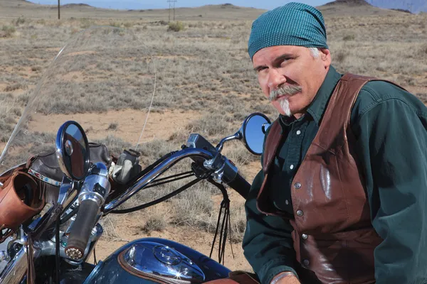 Bello uomo più vecchio appoggiato contro la grande moto all'aperto nel deserto marrone Immagini Stock Royalty Free