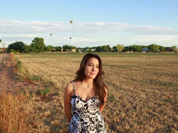 Attractive Hispanic woman standing in open field at sunrise against backdrop of hot air balloons — Stock Photo, Image