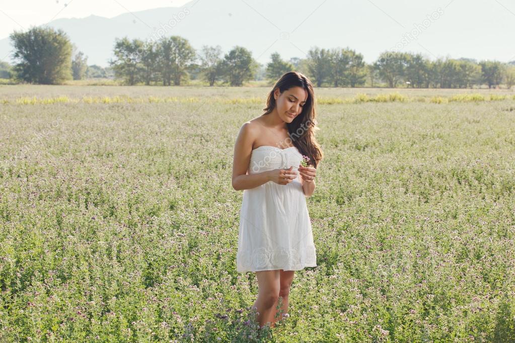 Beautiful Hispanic young woman in white sundress in field of flowers in early morning sunlight