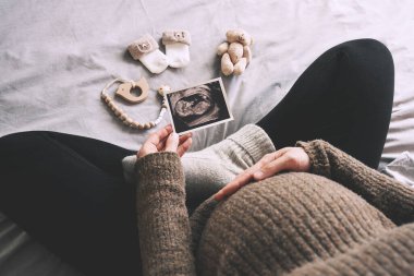 Ultrason görüntüsü olan hamile bir kadının minimalist fotoğrafı, soğuk kış günlerinde yatakta şirin bebek çorapları ve oyuncaklar. Hamile karnı olan hamile bir anne bebek bekliyor. Hamilelik kavramı.