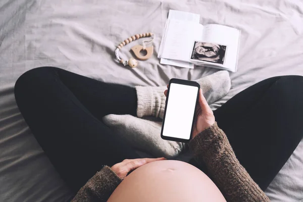 Pregnant woman holding in hands smartphone with empty white display. Close-up pregnant belly of expectant mother with ultrasound photos and medical test reports. Concept of pregnancy, gynecology.