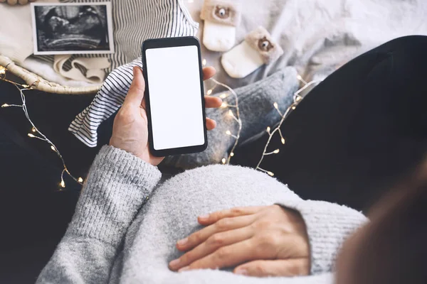 Pregnant woman holding in hands smartphone with empty white display. Close-up pregnant belly of expectant mother with ultrasound photos and medical test reports. Concept of pregnancy, gynecology.