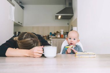 Uykusuz bir geceden sonra modern, yorgun bir anne ve küçük bir çocuk. Bebekli bitkin bir kadın mutfakta kahveyle oturuyor. Bebekle çalışan anne hayatı. Doğum sonrası depresyonu doğum izninde.