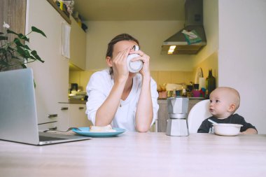 Mutfakta bebekle kahvaltı ederken kahve içmekten yorgun düşmüş genç bir anne. Uykusuz bir geceden sonra serbest çalışan anne ve çocuk. Doğum izninde evde çalışan ya da okuyan bir kadın