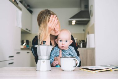 Uykusuz bir geceden sonra modern, yorgun bir anne ve küçük bir çocuk. Bebekli bitkin bir kadın mutfakta kahveyle oturuyor. Bebekle çalışan anne hayatı. Doğum sonrası depresyonu doğum izninde.