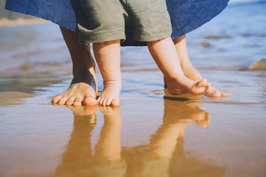 Bebeğin ilk adımları. Anne ve küçük çocuk okyanus kenarında kumsalda yalınayak yürüyor. Doğada mutlu genç bir aile. Anne ve bebek açık deniz sahillerinde oynuyorlar. İnsan ayaklarına yakın çekim.