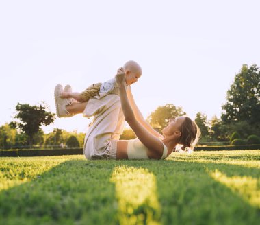 Genç anne ve bebek yazın parkta oynuyorlar. Güzel anne ve çocuk dışarıda. Çocuklu ebeveynler birlikte vakit geçirir. Güneş ışığında mutlu ve sağlıklı bir aile. Çevresel sürdürülebilir yaşam tarzı. Pozitif duygular.