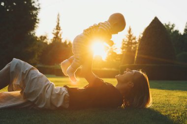 Genç anne ve bebek yazın parkta oynuyorlar. Güzel anne ve çocuk dışarıda. Çocuklu ebeveynler birlikte vakit geçirir. Güneş ışığında mutlu ve sağlıklı bir aile. Çevresel sürdürülebilir yaşam tarzı. Pozitif duygular.