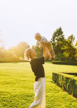 Genç anne ve bebek yazın parkta oynuyorlar. Güzel anne ve çocuk dışarıda. Çocuklu ebeveynler birlikte vakit geçirir. Güneş ışığında mutlu ve sağlıklı bir aile. Çevresel sürdürülebilir yaşam tarzı. Pozitif duygular.