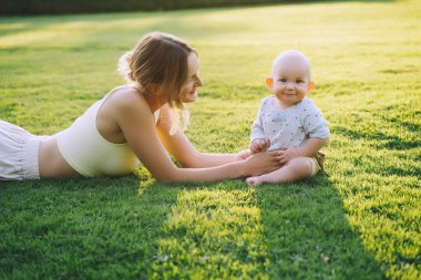 Genç anne ve bebek yazın parkta oynuyorlar. Güzel anne ve çocuk dışarıda. Çocuklu ebeveynler birlikte vakit geçirir. Güneş ışığında mutlu ve sağlıklı bir aile. Çevresel sürdürülebilir yaşam tarzı. Pozitif duygular.
