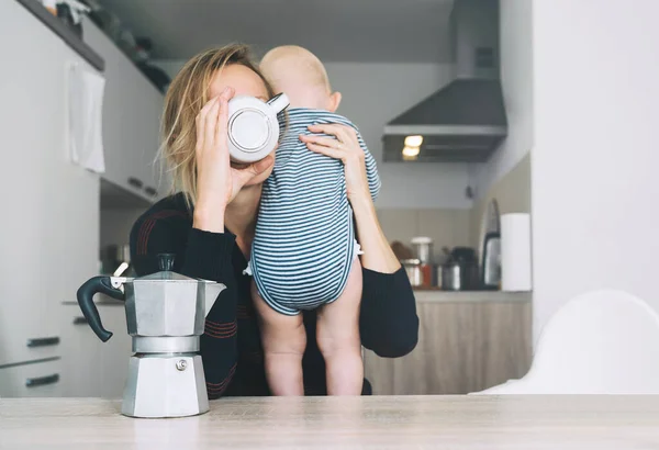 Modern young tired mom and little child after sleepless night. Exhausted woman with baby is sitting with coffee in kitchen. Life of working mother with baby. Postpartum depression on maternity leave.