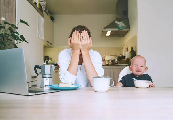 Young millennial mother with coffee fighting tiredness while breakfast with baby in kitchen. Freelancer mom and child after sleepless night. Woman studying or working online at home on maternity leave