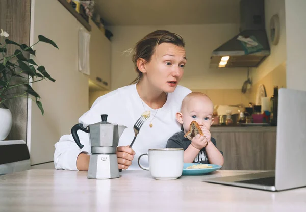 Young millennial mother with coffee fighting tiredness while breakfast with baby in kitchen. Freelancer mom and child after sleepless night. Woman studying or working online at home on maternity leave