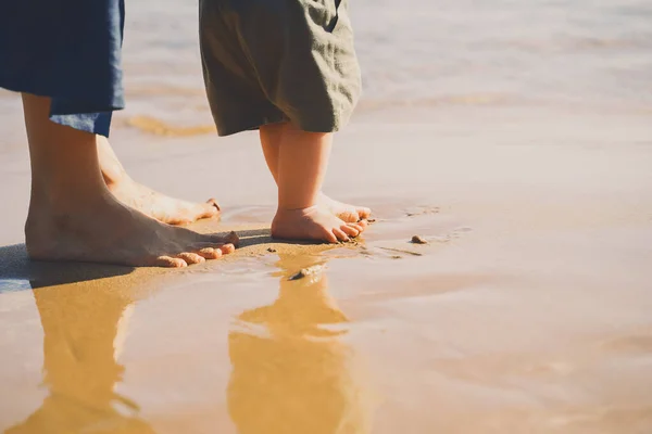 Baby\'s first steps. Mother and small child walking barefoot on beach sand near by ocean. Happy young family in nature. Mother and baby playing outdoors on sea beach. Close-up human feet.