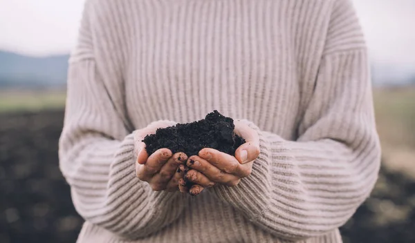 Person holding black soil in hands in background of agricultural field area. Young farmer checking soil. Concept of gardening, ecology environment, sustainable life without plastic. Earth day.