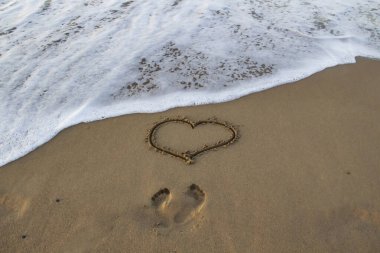 Kalp kumda çizilmiş, sahilde ayak izleri ve önünde deniz köpüğü var..