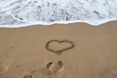 Kalp kumda çizilmiş, sahilde ayak izleri ve önünde deniz köpüğü var..