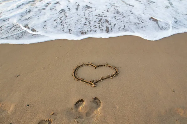 Heart drawn in the sand with footprints on the beach with sea foam in front.