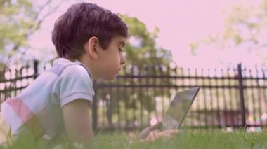 Beyaz küçük çocuk yeşil çimlerin üzerinde oturuyor ve dijital tablet kullanıyor. Bir yaz günü çocuğu tabletle dışarıda tek başına kapat..