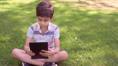 Beyaz küçük çocuk yeşil çimlerin üzerinde oturuyor ve dijital tablet kullanıyor. Bir yaz günü çocuğu tabletle dışarıda tek başına kapat..