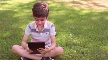 Beyaz küçük çocuk yeşil çimlerin üzerinde oturuyor ve dijital tablet kullanıyor. Bir yaz günü çocuğu tabletle dışarıda tek başına kapat..
