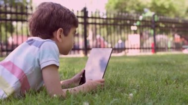 Beyaz küçük çocuk yeşil çimlerin üzerinde oturuyor ve dijital tablet kullanıyor. Bir yaz günü çocuğu tabletle dışarıda tek başına kapat..