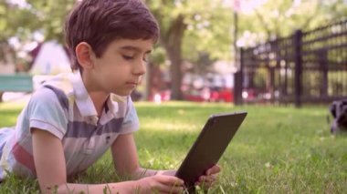 Beyaz küçük çocuk yeşil çimlerin üzerinde oturuyor ve dijital tablet kullanıyor. Bir yaz günü çocuğu tabletle dışarıda tek başına kapat..