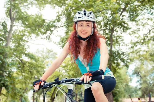 Menina ruiva bonita montando e pedalando uma bicicleta em um parque da cidade — Fotografia de Stock