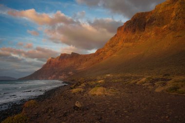 Famara Sahili yakınlarındaki kayalıklarda gün batımı, Lanzarote, İspanya. Mavi gökyüzündeki bazı bulutlar. Son gün ışığı dağın tepelerinde parlıyor..