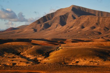 Costa de Papagayo, Lanzarote, İspanya 'da. Gün batımında Los Ajaches sıradağları. Mavi gökyüzünde bulutlarla güneşli bir akşam, altın saat. Ortada yürüyen iki kişi..
