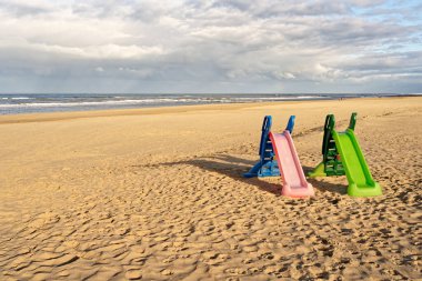 Two playground slides on an almost empty beach, one green and one pink. North Holland dune reserve, Egmond aan Zee, Netherlands.