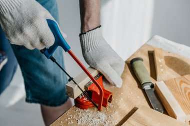Ev ve bahçe için el yapımı raf üretimi için malzeme hazırlama. İş eldivenli bir adam küçük bir tahta parçasını testereyle kesiyor. ÇEVİR.