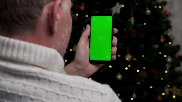 白いセーターに身を包んだ中年のおじさんが クリスマスツリーを背景に緑色の画面をしたスマートフォンを持っています クロマキーで携帯電話を閉じる — ストック動画