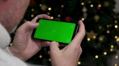 4K. Orta yaşlı bir adam akıllı telefonuyla oyun oynuyor. Kromakey ile yeşil ekranda, ekranda parmaklarına basıyor. Yılbaşı ağacının arka planında parlayan bir çelenkle.