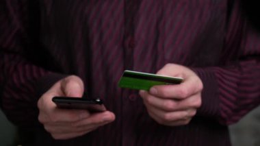 4K yakın plan, elinde kredi kartı, akıllı telefon ve internetten alışveriş yapan bordo gömlekli bir adamın elleri. Bankacılık, online alışveriş ve yaşam tarzı konsepti. Modern teknolojiler