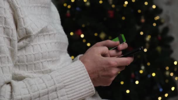 4Kクリスマスツリーの近くに座っている男は クレジットカードとスマートフォンを持ち 即時送金 オンライン購入を行っています クリスマスセール インターネットでの贈り物の購入 オンライン銀行 — ストック動画