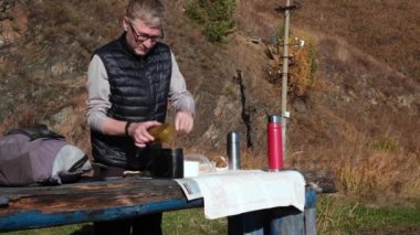 Sonbaharda doğada piknik. Ahşap bir masada duran bir adam, sıcak şehriye çorbasını bir kaba dolduruyor. Circum-Baykal Demiryolu boyunca yürüyüş. Yerel seyahatler ve hafta sonu gezileri. 4K.