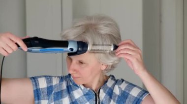 4K. An attractive middle-aged woman independently dries and styles her short gray hair with a hairdryer at home. Close-up.