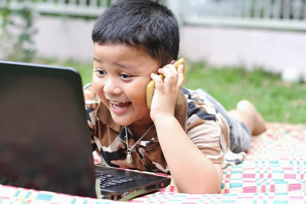 The boys are playing a laptop and talking on the phone. Happily. — Stock Photo, Image