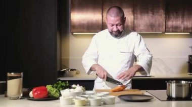 Mutfakta önlüklü bir adam ev yapımı öğle yemeği hazırlıyor. Aşçı lezzetli bir yemek için taze havuçları keskin bir bıçakla keser. Malzemelerin hazırlanması
