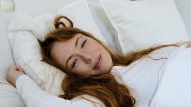 Parlak beyaz yatak odasındaki kızıl saçlı kız beyaz yastıkta rahat bir yatakta yatıyor. Güzel uyuyan dişi sabah uyanır ve gözlerini açar, kendini battaniyeye sarar..