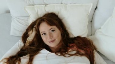 Günaydın hafta sonu. Mutlu genç bir kadın, mutlu bir şekilde yatak odasında uyanıyor, kafası yastığın üzerinde uzanıyor, gülümsüyor ve zevkle esniyor. Üst görünüm.