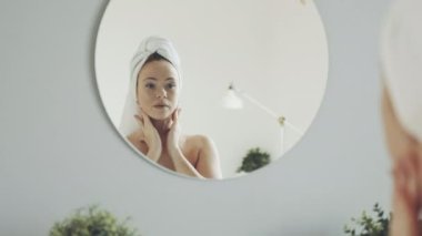 Genç, mutlu ve çekici bir kadın sabah banyo aynasında yansımaya bakar, duş aldıktan sonra kafasında havlu olan sağlıklı genç bayan yüzüne nemlendirici sürer ve cildine masaj yapar..