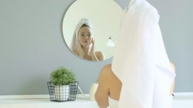 Kafasında beyaz havlu olan kadın, banyoda duruyor, kavanozdan nemlendiriciyi alıyor ve yüzüne masaj yapıyor. Kız cilt ve vücut bakımı için kozmetik kullanıyor