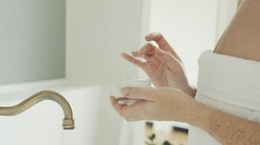 Kafasında beyaz havlu olan kadın, banyoda duruyor, kavanozdan nemlendiriciyi alıyor ve yüzüne masaj yapıyor. Kız cilt ve vücut bakımı için kozmetik kullanıyor