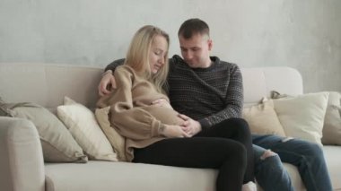 Genç ve mutlu bir aile bebek bekliyor. Kanepede oturup sarılıyorlar. Bekleyen neşeli ebeveynler kadının karnını okşar ve mutlulukla gülerler.