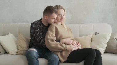 Genç ve mutlu bir aile bebek bekliyor. Kanepede oturup sarılıyorlar. Neşeli, hamile ebeveynler kadının karnını okşar ve mutlulukla gülerler. Babam hamile anneye bakıyor.