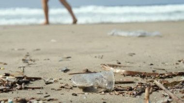 Tek kullanımlık plastik bardak deniz kıyısındaki ekosisteme atılmış ve arka planda yürüyen insanlar, kirli doğa ortamı. 4k